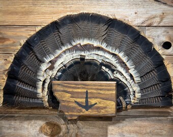 May include: A fan of turkey feathers with black, brown, and white hues, mounted on a wooden surface. A wooden plaque in the shape of Montana with a black arrow is centered below the feathers.