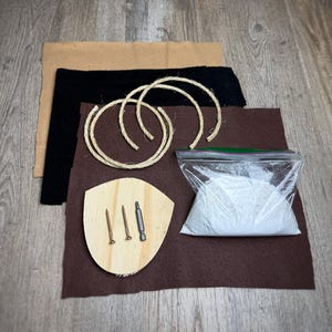 May include: A DIY kit for making a macrame wall hanging. The kit includes a wooden plaque, two pieces of rope, a bag of white powder, two screws, a screwdriver, and three pieces of brown felt.