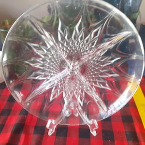 Puede incluir: Un plato de servir de vidrio transparente, redondo y dividido, con un diseño de estrella en el centro. El plato está sostenido por un soporte de plástico transparente y se encuentra sobre un mantel a cuadros rojos y negros.