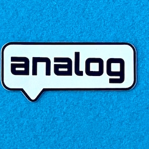 Op de afbeelding: Witte sticker met een zwarte rand in de vorm van een spreekballon. Het woord "analog" is in zwarte letters op de sticker gedrukt.