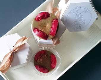 May include: Two white hexagonal boxes with gold ribbon and a red and gold wax melt in a clear container. The wax melt is shaped like a cupcake and has a gold glitter topping. The box has a label that says "Cheer Cherry Muffin" and "Wolfhaven Scents".