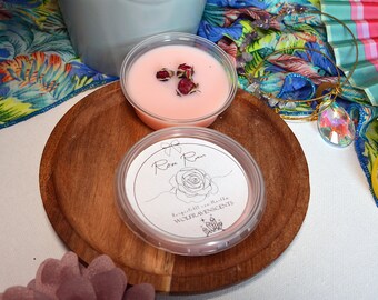 May include: A small pink wax melt in a clear container with a white label that says "Rose Rain" and "Handcrafted with Love WOLF RAVEN SCENTS". The wax melt is on a wooden surface with dried rose buds.