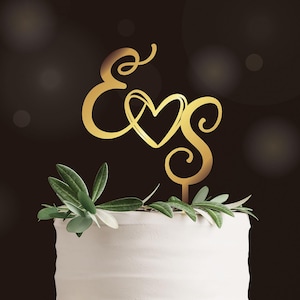 May include: A gold cake topper with the letters "E" and "S" intertwined with a heart design. The topper sits atop a white cake, decorated with green foliage. The background is a dark brown with soft, blurred lights.