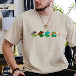 May include: An ivory natural colored t-shirt featuring a graphic of four circular characters in green, each wearing a different colored mask in purple, red, blue, and orange, with the last character facing a small triangular slice of pizza.