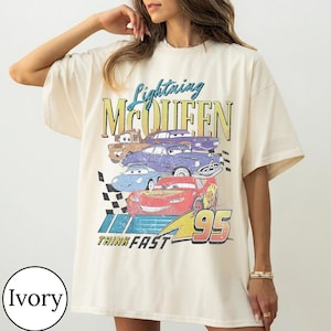 May include: Ivory-colored t-shirt featuring a graphic of Lightning McQueen and other Cars characters. The design includes the text "Lightning McQueen", "Think Fast", and the number "95". The shirt has a vintage, distressed look.