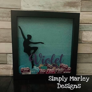 May include: A framed shadow box featuring a black silhouette of a ballerina on a teal background. The name "Violet" is written in purple script. The box includes various "PAC Gold" and "Platinum" stickers. The text "Simply Marley Designs" is at the bottom.