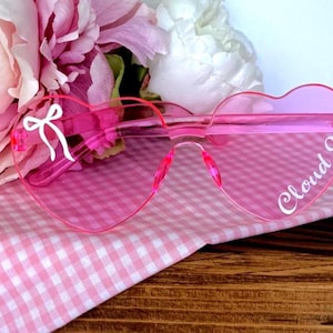 May include: Pink heart-shaped sunglasses with the text "Cloud 9" and a white bow design. The sunglasses are set against a pink gingham cloth and a backdrop of pink and white flowers.