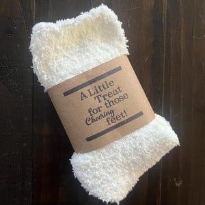 May include: A white, fluffy, spa-like foot scrub wrapped in brown paper with the text "A Little Treat for those Cheering Feet!"