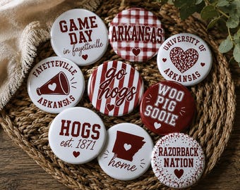 May include: A collection of nine round pinback buttons featuring University of Arkansas themes in red and white. Designs include text such as Game Day in Fayetteville, Go Hogs, Woo Pig Sooie, Razorback Nation, and Hogs est. 1871, with graphics of a megaphone, a heart, and the state of Arkansas.
