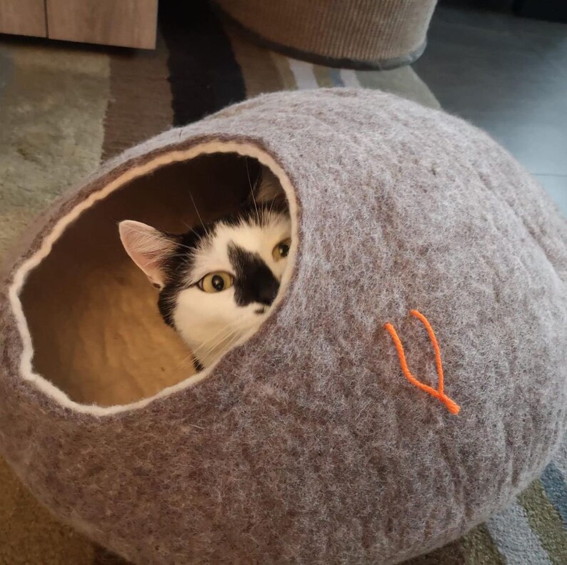 May include: A black and white cat peeks out of a gray felt cat bed. The bed has a round shape and a small opening for the cat to enter. The bed is made of a soft, fuzzy material.