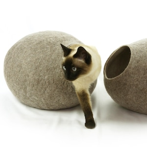 May include: A brown felt cat bed shaped like an egg with a cat peeking out of the opening. The cat is a Siamese with brown and white fur.
