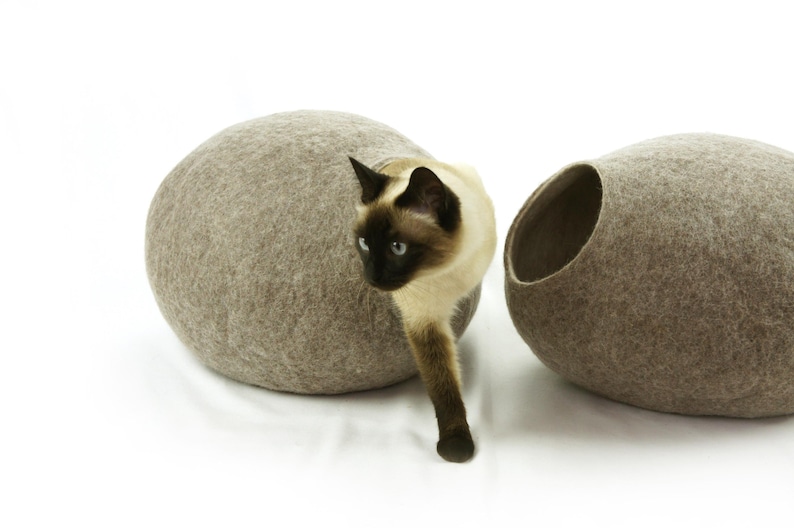 May include: A brown felt cat bed shaped like an egg with a cat peeking out of the opening. The cat is a Siamese with brown and white fur.