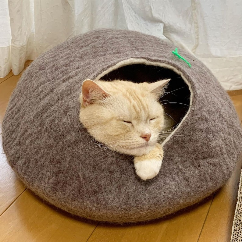 May include: A light brown, round cat bed made of felt with a cream colored interior. A light orange tabby cat is sleeping inside the bed with its paws tucked under its chin.