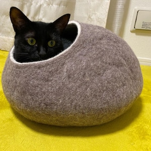 May include: A black cat peeks out of a brown felt cat bed shaped like a rounded oval.