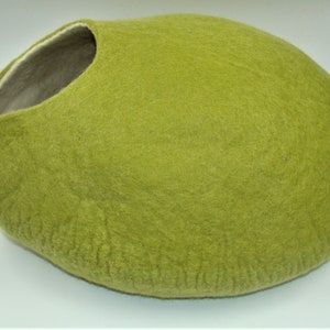 Puede incluir: Una cama para gatos de fieltro verde con forma ovalada. La cama tiene un forro de fieltro blanco alrededor de la abertura.