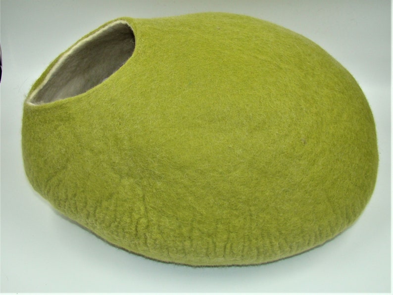 Puede incluir: Una cama para gatos de fieltro verde con forma ovalada. La cama tiene un forro de fieltro blanco alrededor de la abertura.