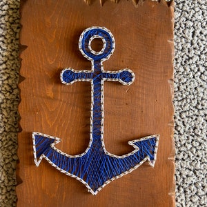 May include: A blue and white string art anchor on a brown wooden background.