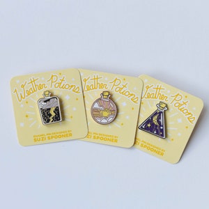 May include: Three enamel pins featuring weather potions in glass bottles. The first pin shows a bottle with a stormy sky, lightning, and clouds. The second pin shows a bottle with a sun, clouds, and a rainbow. The third pin shows a bottle with a crescent moon, stars, and a purple sky. Each pin has the text "Weather Potions" and "Enamel Pin Designed by Suzi Spooner".