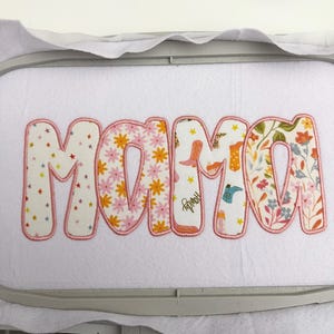 May include: Embroidered fabric applique spelling "MAMA" in bubble letters. The letters are filled with floral and cowboy boot patterns, outlined in pink. The applique is on a white background.