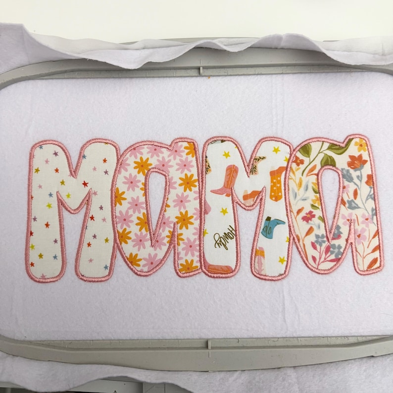 May include: Embroidered fabric applique spelling "MAMA" in bubble letters. The letters are filled with floral and cowboy boot patterns, outlined in pink. The applique is on a white background.