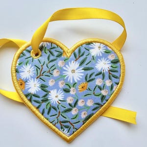 May include: A heart-shaped fabric ornament featuring a light blue background with a pattern of white and yellow flowers and green leaves. The edges are finished with yellow embroidery stitching and a yellow satin ribbon loop for hanging.