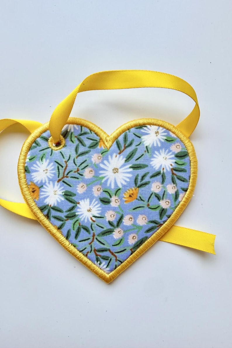 May include: A heart-shaped fabric ornament featuring a light blue background with a pattern of white and yellow flowers and green leaves. The edges are finished with yellow embroidery stitching and a yellow satin ribbon loop for hanging.