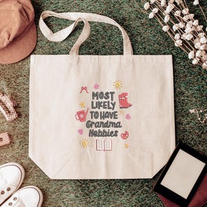 May include: Beige tote bag with the text "MOST LIKELY TO HAVE Grandma Hobbies" in a colorful design. The bag has two handles and is surrounded by a brown cap, white sneakers, and other craft-related items.