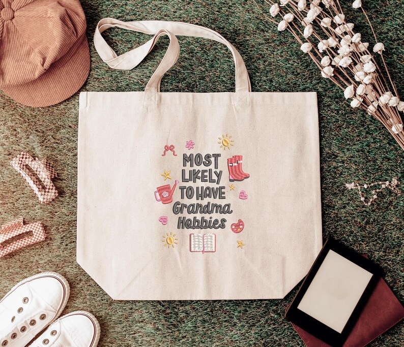May include: Beige tote bag with the text "MOST LIKELY TO HAVE Grandma Hobbies" in a colorful design. The bag has two handles and is surrounded by a brown cap, white sneakers, and other craft-related items.