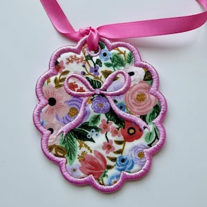 May include: A scalloped, circular fabric ornament featuring a white background with a colorful floral print of pink, red, blue, and purple flowers and green leaves. The edge is finished with pink embroidery and a matching pink ribbon loop.
