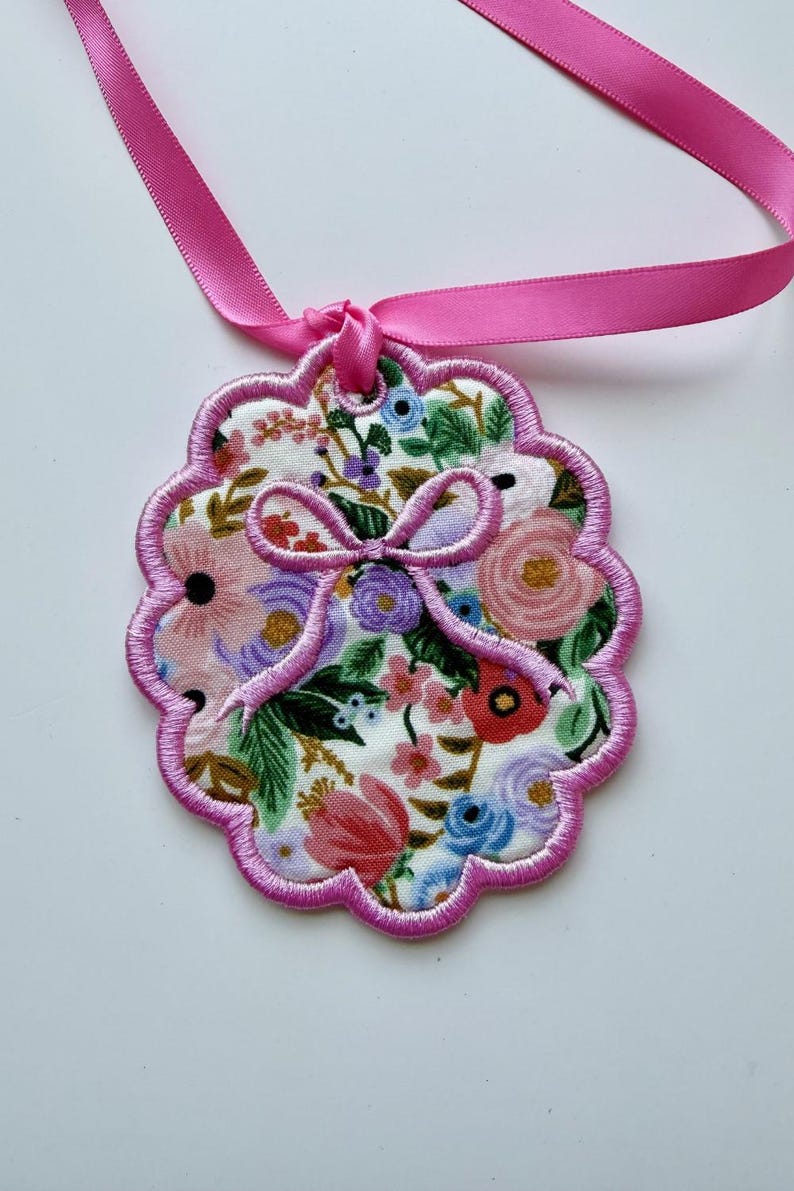 May include: A scalloped, circular fabric ornament featuring a white background with a colorful floral print of pink, red, blue, and purple flowers and green leaves. The edge is finished with pink embroidery and a matching pink ribbon loop.