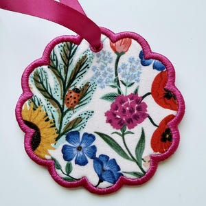 May include: A scalloped, circular fabric ornament featuring a white background with a colorful floral print including yellow sunflowers, red poppies, blue flowers, and a ladybug. The edge is finished with bright pink embroidery and a pink ribbon.