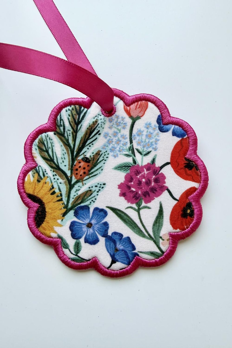 May include: A scalloped, circular fabric ornament featuring a white background with a colorful floral print including yellow sunflowers, red poppies, blue flowers, and a ladybug. The edge is finished with bright pink embroidery and a pink ribbon.