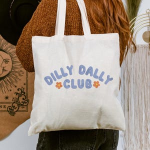 May include: Cream-colored tote bag with the words "DILLY DALLY CLUB" in blue, stylized text. The word "CLUB" is in a smaller font size. Orange flower graphics are on either side of the word "CLUB". The bag has two white handles.