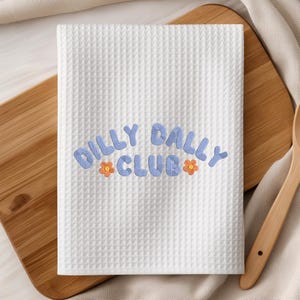 May include: White waffle-weave kitchen towel with the embroidered text "Dilly Dally Club" in light blue, accented with orange flower details. The towel is rectangular and folded, suggesting it's ready for use.