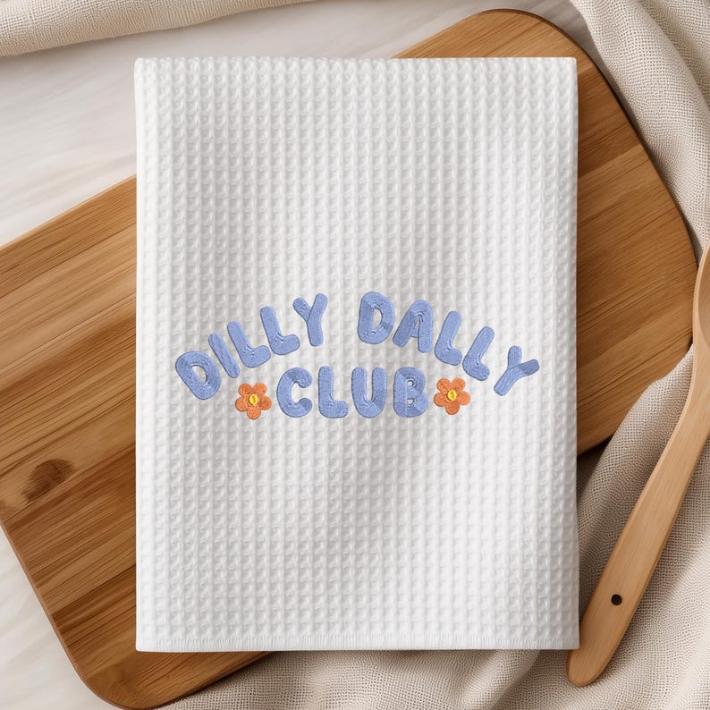 May include: White waffle-weave kitchen towel with the embroidered text "Dilly Dally Club" in light blue, accented with orange flower details. The towel is rectangular and folded, suggesting it's ready for use.