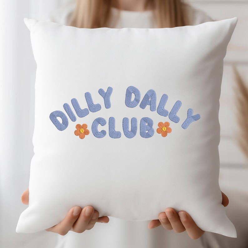May include: White square pillow with the embroidered text "DILLY DALLY CLUB" in blue, with orange flower accents. The pillow is held by a person.