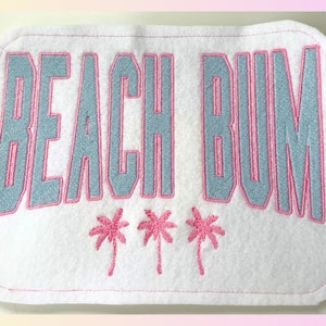 May include: White fabric patch with pink stitching. The patch says "Beach Bum" in blue with a pink outline. There are three pink palm trees below the text.
