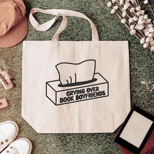 Puede incluir: Bolso tote beige con texto negro &laquo;Crying Over Book Boyfriends&raquo; y un gr&aacute;fico de caja de pa&ntilde;uelos. El bolso tiene forma rectangular y dos asas. Otros art&iacute;culos incluyen una gorra, zapatos y pinzas para el pelo.