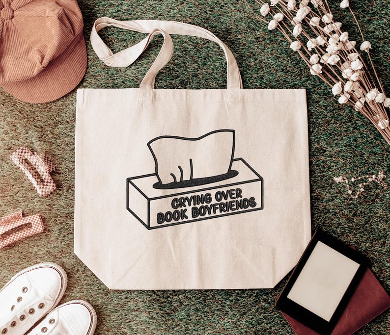 Puede incluir: Bolso tote beige con texto negro &laquo;Crying Over Book Boyfriends&raquo; y un gr&aacute;fico de caja de pa&ntilde;uelos. El bolso tiene forma rectangular y dos asas. Otros art&iacute;culos incluyen una gorra, zapatos y pinzas para el pelo.