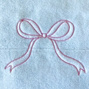 May include: Close-up of a white textile with a pink embroidered bow design. The bow has two loops and two flowing ribbons. The embroidery thread has a subtle sheen, adding a touch of elegance to the simple design.
