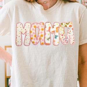May include: Cream-colored t-shirt with the word "MAMA" in a colorful, floral design. The letters are outlined in pink and filled with patterns of stars, flowers, and cowboy boots. A gold butterfly necklace is visible.