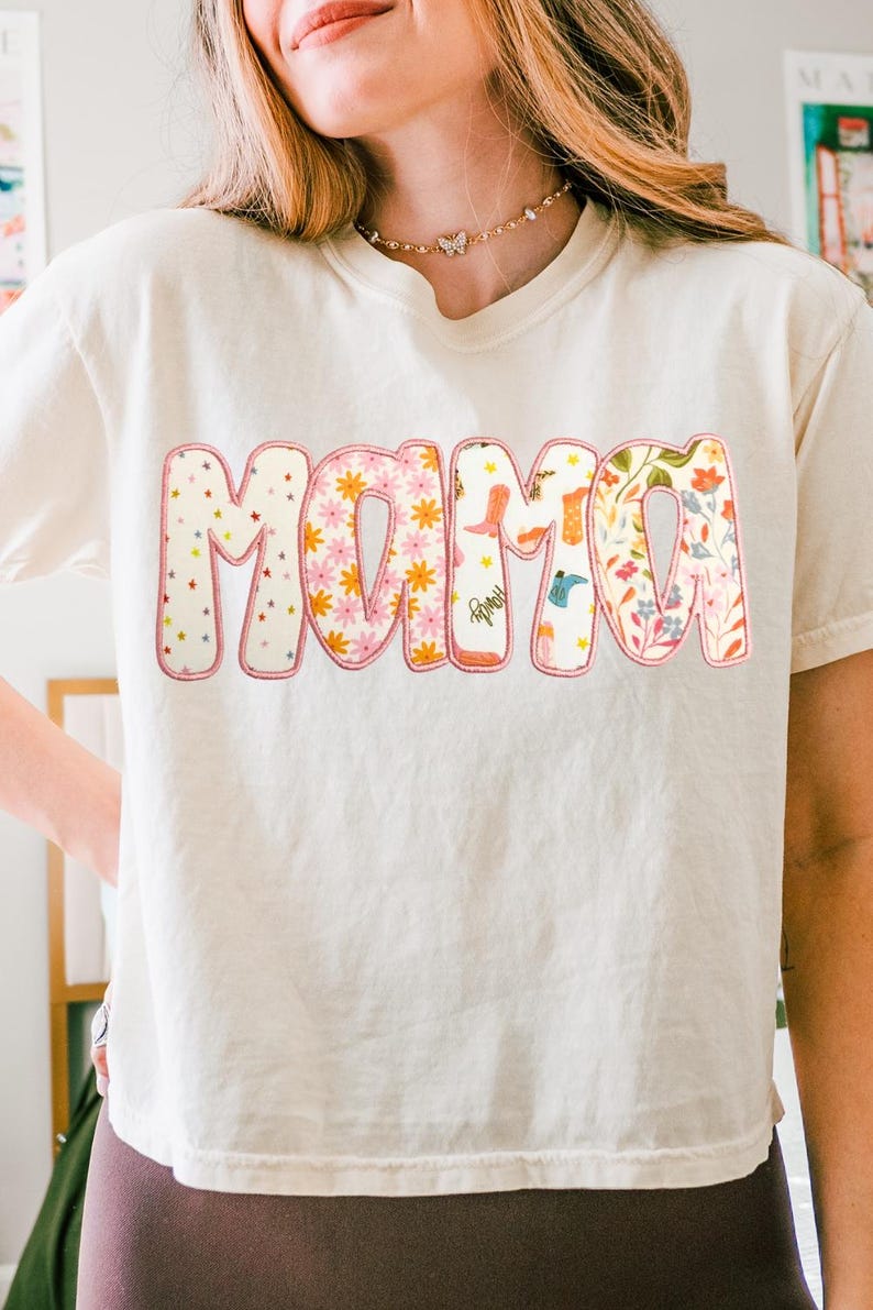 May include: Cream-colored t-shirt with the word "MAMA" in a colorful, floral design. The letters are outlined in pink and filled with patterns of stars, flowers, and cowboy boots. A gold butterfly necklace is visible.