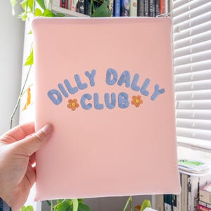 May include: A light pink zippered pouch with the words "DILLY DALLY CLUB" in blue, with orange flower accents. The pouch is held in a person's hand, with a white zipper and a light pink interior.
