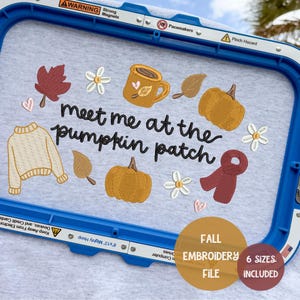 May include: A light gray fabric in a blue embroidery hoop featuring the embroidered text "meet me at the pumpkin patch" surrounded by icons of a beige sweater, two orange pumpkins, a brown mug, a red scarf, brown leaves, and white flowers.