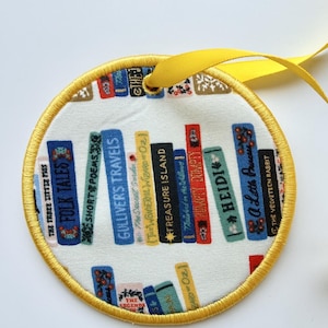 May include: A circular fabric ornament featuring a print of colorful book spines on a white background. The books include titles like Gulliver's Travels and Treasure Island. The ornament has a yellow stitched border and a yellow ribbon for hanging.
