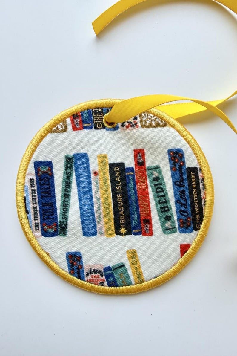 May include: A circular fabric ornament featuring a print of colorful book spines on a white background. The books include titles like Gulliver's Travels and Treasure Island. The ornament has a yellow stitched border and a yellow ribbon for hanging.