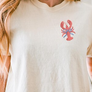 May include: Cream-colored t-shirt featuring a red embroidered lobster design with a blue bow. The shirt has short sleeves and a relaxed fit. The lobster is centered on the chest. The overall style is casual and summery.