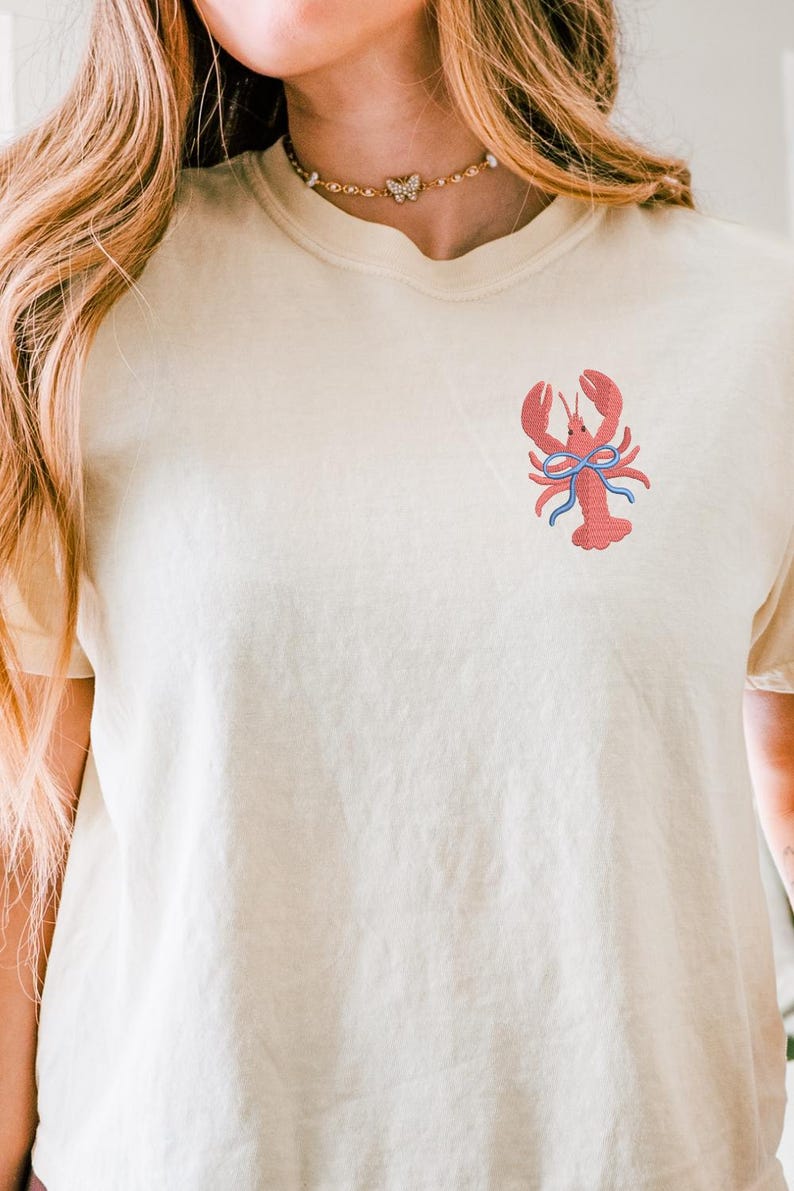 May include: Cream-colored t-shirt featuring a red embroidered lobster design with a blue bow. The shirt has short sleeves and a relaxed fit. The lobster is centered on the chest. The overall style is casual and summery.
