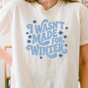 May include: Cream-colored t-shirt with the phrase "I Wasn't Made For Winter" in light blue, surrounded by blue starbursts. The shirt is a casual, short-sleeved style.