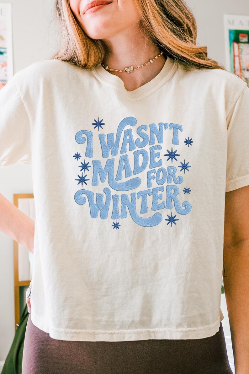 May include: Cream-colored t-shirt with the phrase "I Wasn't Made For Winter" in light blue, surrounded by blue starbursts. The shirt is a casual, short-sleeved style.